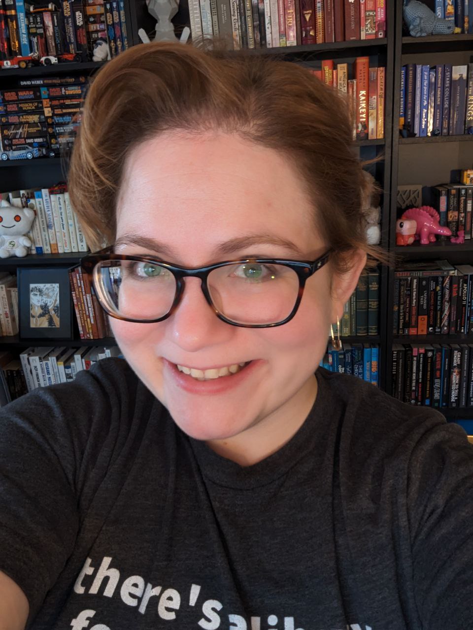 A picture of a woman in her early thirties looking at the camera. Her reddish hair is neatly coiffed and she's smiling at the camera in front of bookshelves.